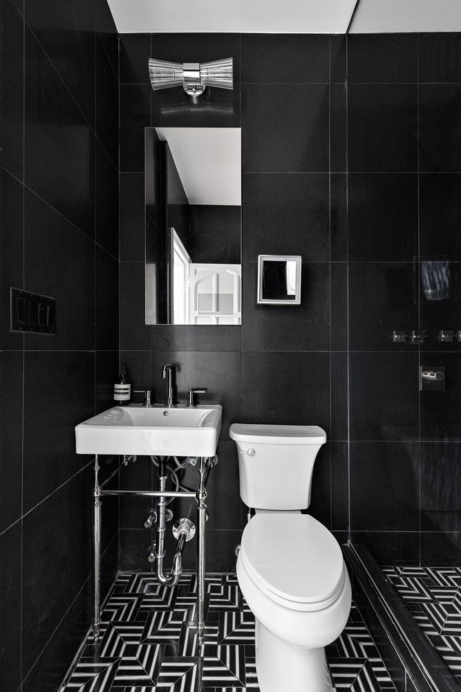 This is a modern bathroom featuring black tiled walls and a striking black and white patterned floor. A white pedestal sink with chrome fixtures sits next to a white toilet. A mirror and modern light fixture complete the space, creating a sleek and stylish look.