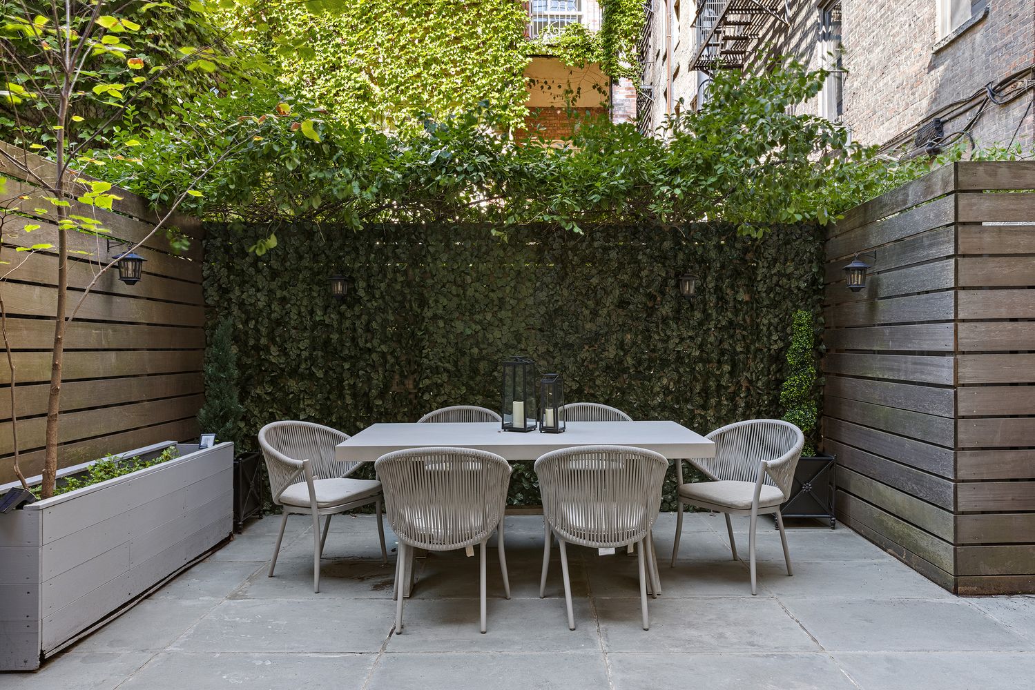This is an exterior shot of a charming patio area, featuring a dining set with six chairs and a rectangular table. The patio is enclosed by wooden fencing and a lush green wall of ivy, creating a private and serene outdoor space. The flooring is made of large stone tiles, and the overall impression is one of tranquility and sophisticated outdoor living.