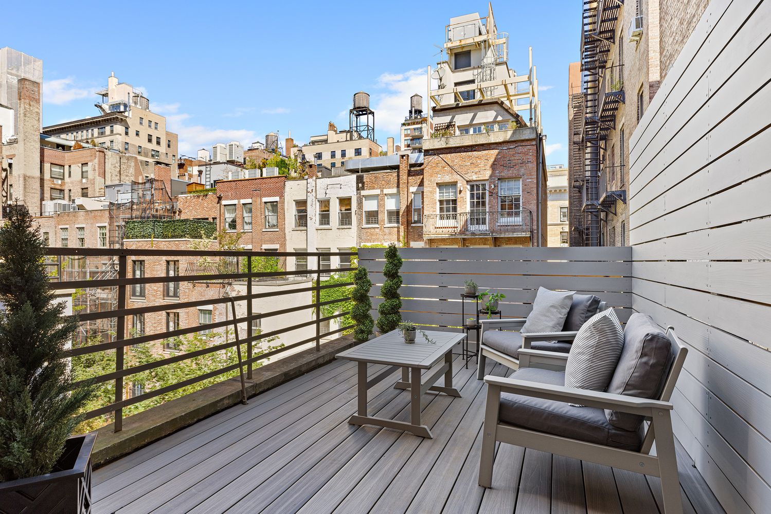 This image showcases a charming outdoor balcony or deck area, furnished with comfortable seating and a small table, creating an inviting space for relaxation. The deck is enclosed by a railing and a privacy screen, offering a sense of seclusion while still providing views of the surrounding cityscape. The overall impression is one of urban tranquility and stylish outdoor living.