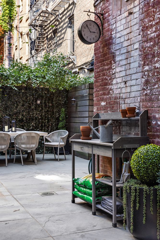 This image showcases a charming outdoor patio or garden area, featuring a potting bench with gardening supplies, a dining table with chairs, and lush greenery providing privacy. The brick wall and vintage clock add character, creating a cozy and inviting atmosphere. The space is well-maintained and designed for relaxation and outdoor enjoyment.