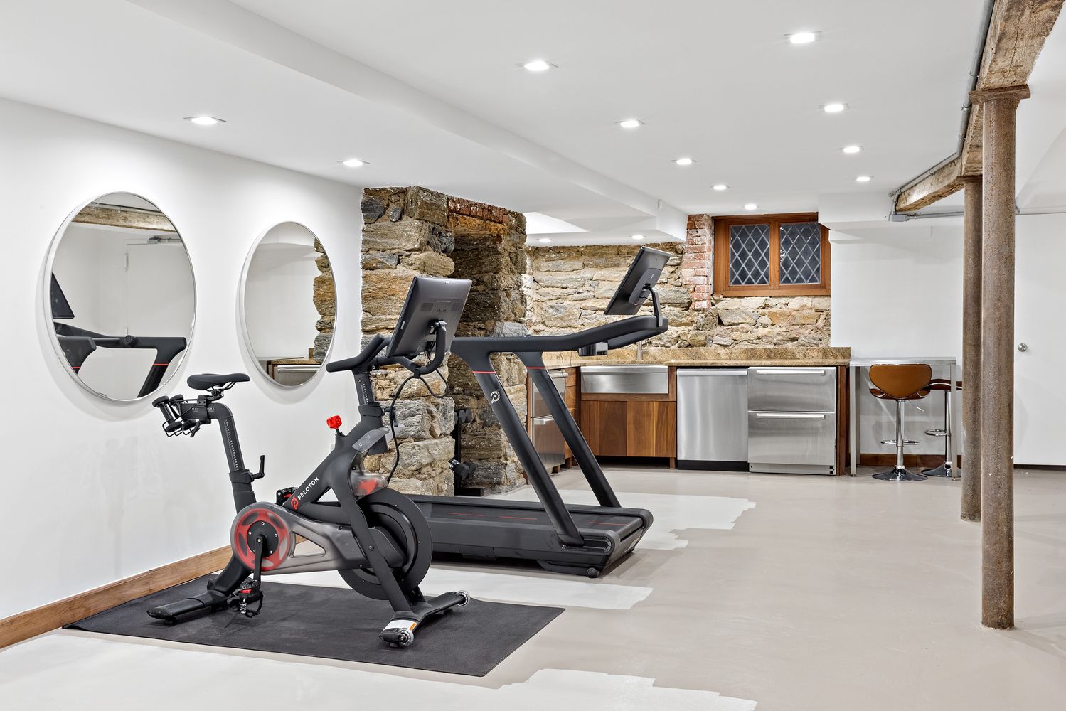 This is a well-appointed home gym featuring a Peloton bike and a treadmill. The space has a unique design with exposed stone walls and modern fixtures, including round mirrors and recessed lighting. The floor is a light neutral color, and the overall impression is clean and functional.