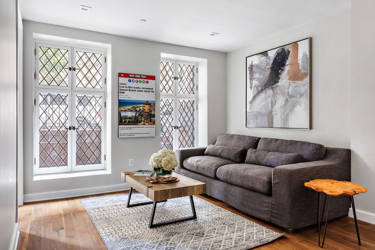 This is a cozy living room featuring a comfortable gray sofa, a wooden coffee table with a floral arrangement, and an abstract art piece hanging on the wall. The room is well-lit by natural light coming through the windows, which have a diamond pattern, and a New York Post article is displayed on the wall between the windows. The hardwood floors and patterned rug add warmth and texture to the space.