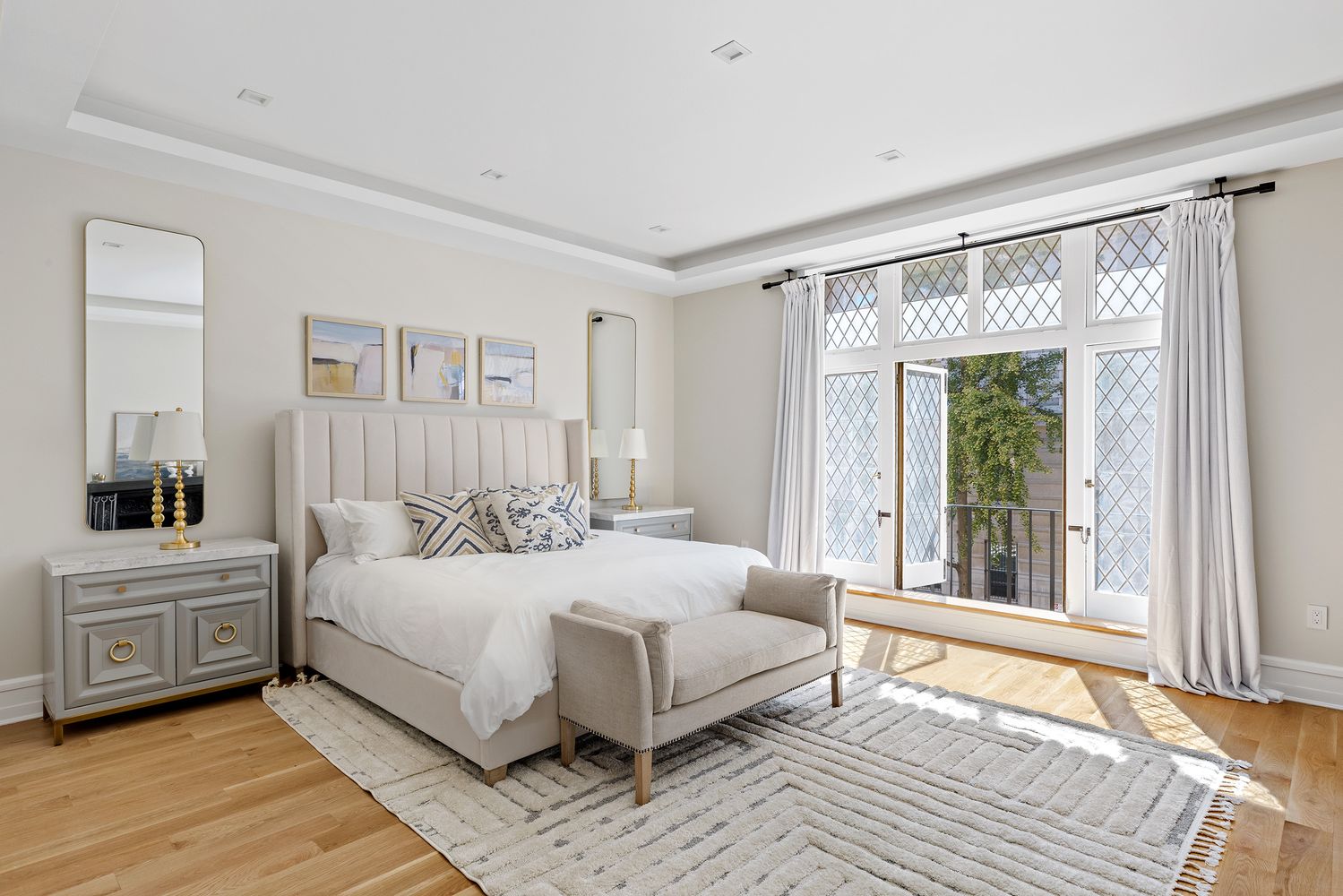 This is a bright and airy primary bedroom featuring a large bed with a neutral-toned headboard, complemented by decorative pillows and a cozy bench at the foot. The room is well-lit with natural light streaming through large windows, enhanced by light-colored walls and hardwood floors. A stylish rug adds texture, while elegant side tables and artwork contribute to the room's sophisticated and inviting atmosphere.