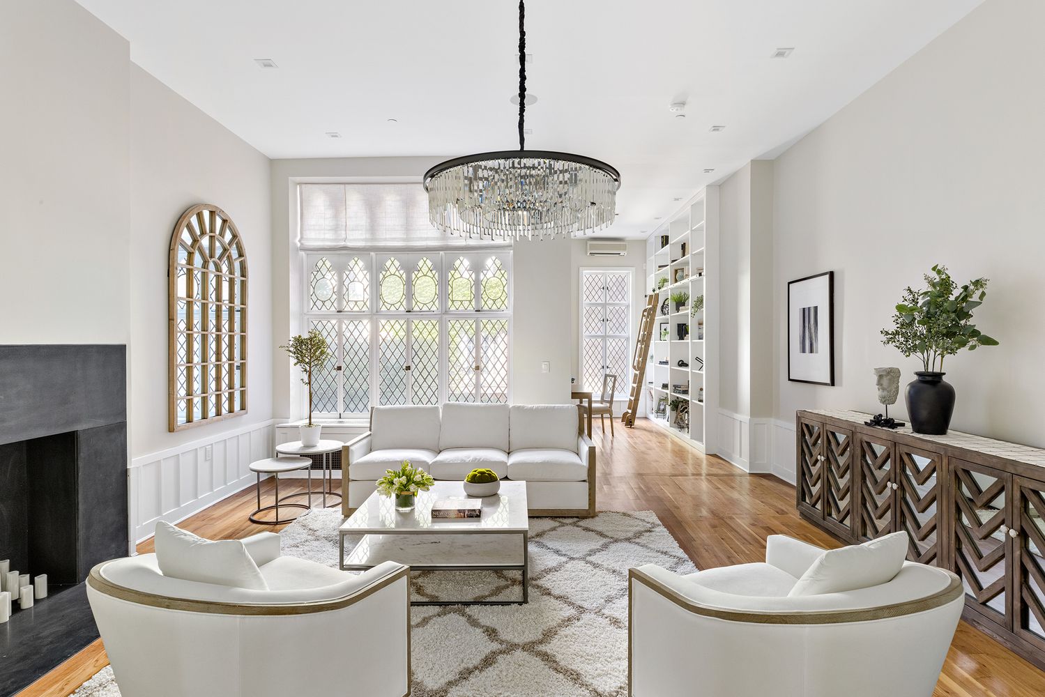 This is a bright and elegant living room featuring white walls, hardwood floors, and a large window with decorative glass. The room is furnished with a white sofa, two armchairs, a marble coffee table, and a decorative console table with a vase of greenery. A large chandelier hangs from the ceiling, adding a touch of sophistication.