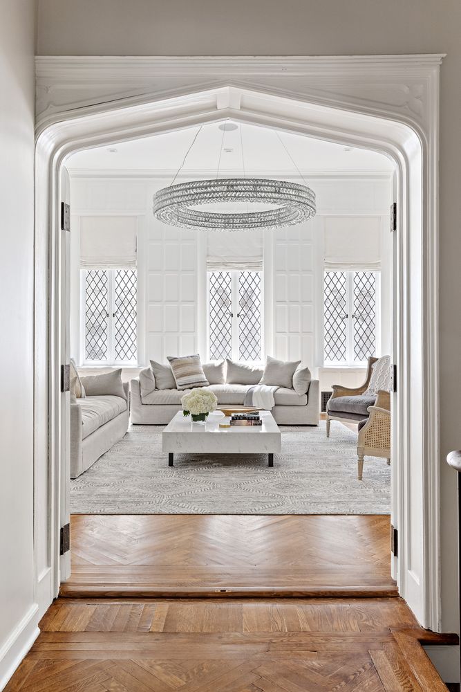 This interior shot showcases a bright and elegant living room, viewed through an arched doorway. The room features a neutral color palette, with comfortable sofas, a marble coffee table, and a striking circular chandelier. Three large windows with diamond-patterned panes provide ample natural light, enhancing the room's sophisticated and inviting atmosphere.