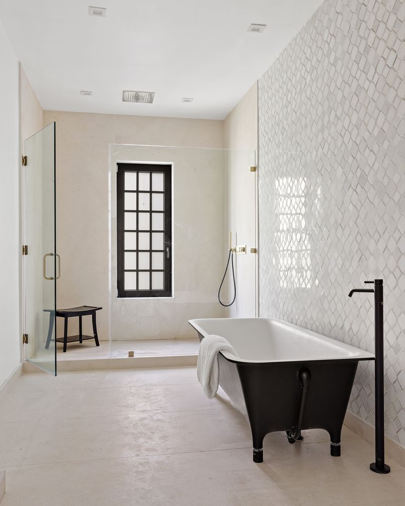 This primary bathroom features a freestanding black bathtub with a floor-mounted faucet, set against a backdrop of patterned tile. A glass-enclosed shower with a black-framed window adds a modern touch, while the neutral color palette creates a serene and luxurious atmosphere. The perspective is from the doorway, showcasing the depth and layout of the space.
