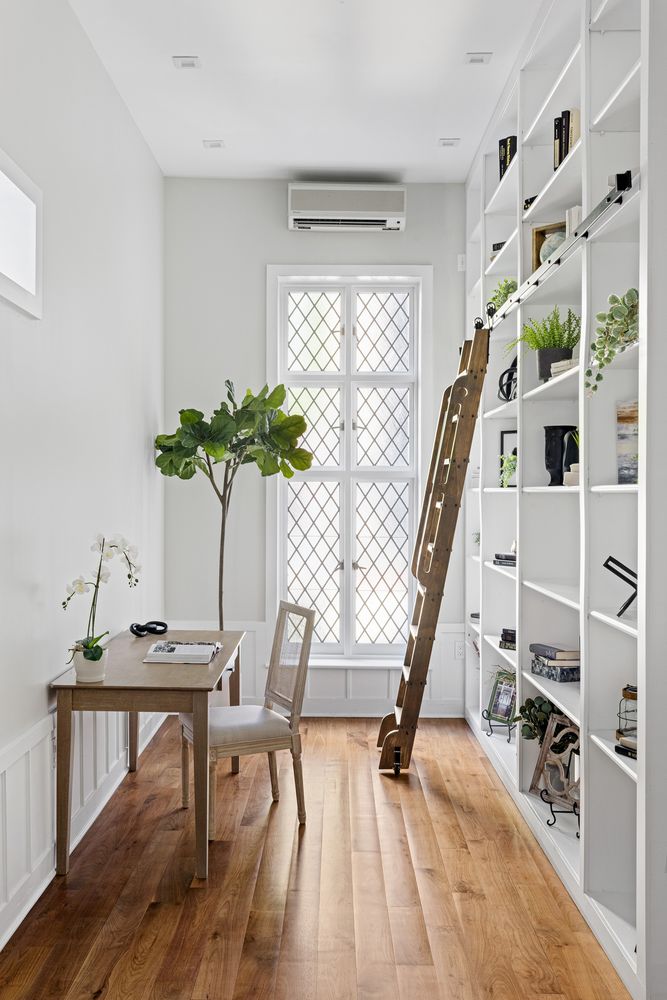 This is an interior shot of a bright and airy home office or study. The room features a wooden desk with a chair, a large window with diamond-patterned panes, and a tall, built-in bookshelf with a rolling ladder. The hardwood floors and neutral color palette create a warm and inviting atmosphere, perfect for a productive workspace.