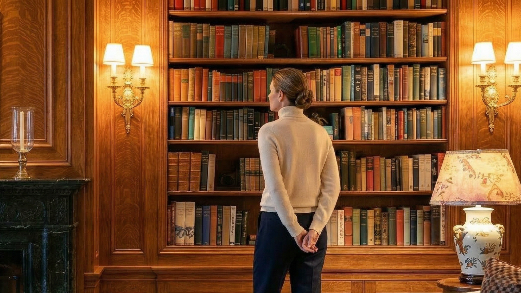 This sophisticated study features floor-to-ceiling wooden built-in bookshelves filled with an extensive collection of volumes, providing an elegant and scholarly ambiance. The space is highlighted by warm decorative wall sconces, a traditional lamp with a patterned shade, and rich wood paneling that enhances the room's timeless, luxurious character.