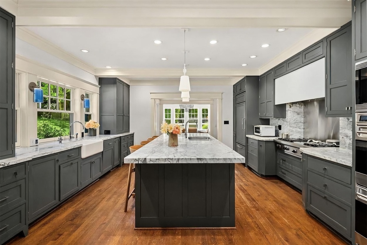 This sophisticated kitchen features dark gray cabinetry accented by white marble countertops and a matching marble backsplash. A prominent central island with bar seating serves as a functional focal point, while rich hardwood floors and recessed lighting enhance the warm and elegant atmosphere. The space is well-equipped with integrated modern appliances and overlooks a bright, windowed breakfast area.