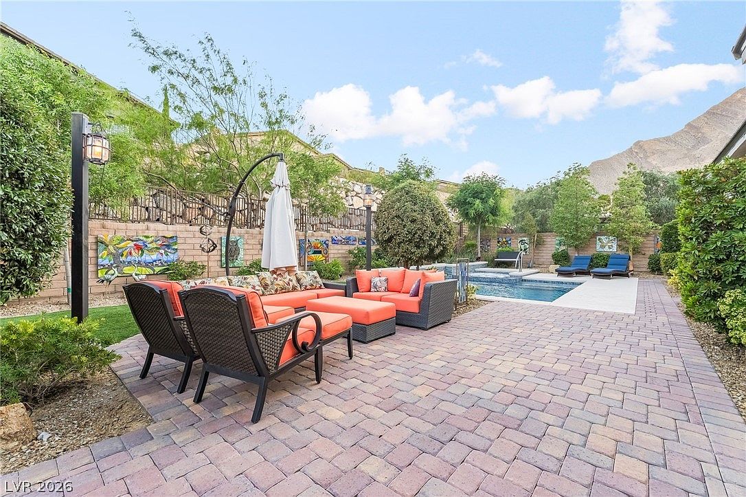 This spacious backyard patio features high-quality stone pavers and a comfortable outdoor sectional seating area with bright orange cushions, perfect for relaxation and entertaining. The space transitions seamlessly into a well-maintained pool and spa area, framed by landscaped greenery and a decorative retaining wall against a mountainous backdrop. The overall design offers a private, resort-like atmosphere ideal for luxury outdoor living.