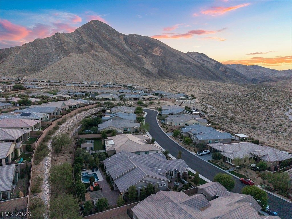 This stunning aerial view captures a desert residential community nestled at the base of a majestic mountain range. The neighborhood features modern homes with tile roofs, manicured landscaping, and select properties with private swimming pools. The golden hour lighting highlights the dramatic landscape and the picturesque, high-end suburban atmosphere.