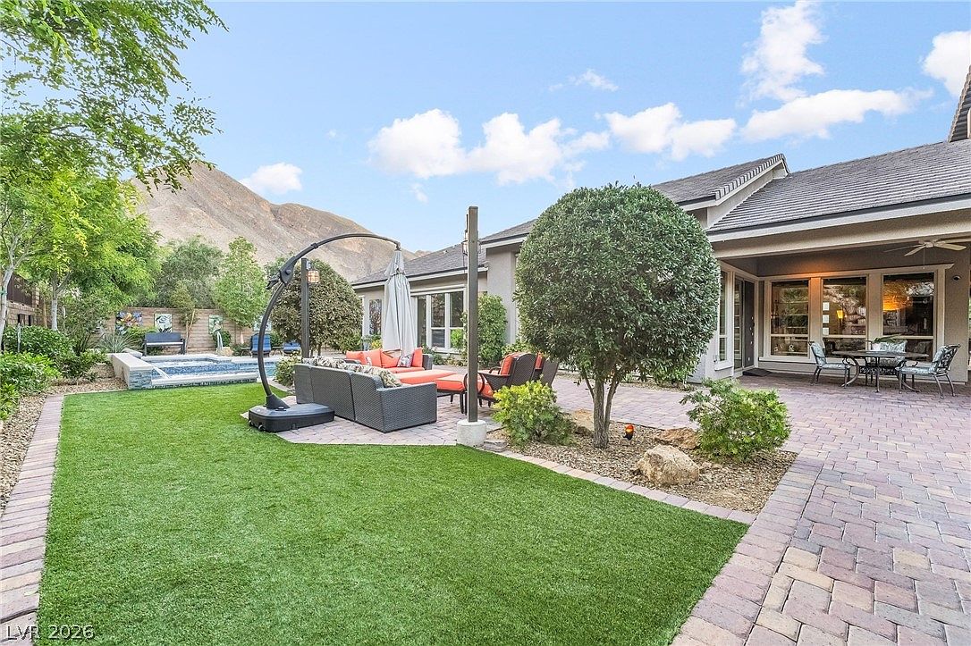 This spacious backyard features a beautifully paved patio area perfect for outdoor dining and lounging. It includes a green artificial turf lawn, an inviting hot tub, and mature landscaping set against a picturesque mountain backdrop. The home's rear exterior provides seamless access to this private, resort-style retreat.