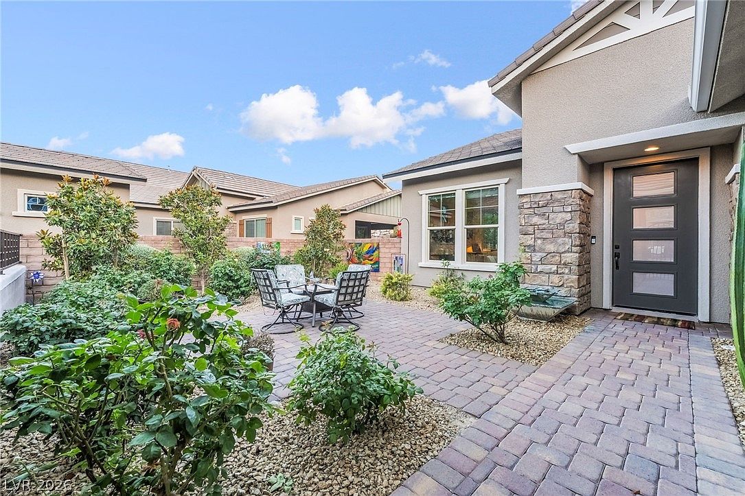 This appealing outdoor living space features a well-maintained paver patio set against a home with stucco siding and stone architectural details. The area is landscaped with various shrubs and desert-style gravel, creating a tranquil garden atmosphere perfect for relaxation. A modern, dark-framed glass front door adds a sophisticated touch, seamlessly connecting the interior to this inviting private courtyard.