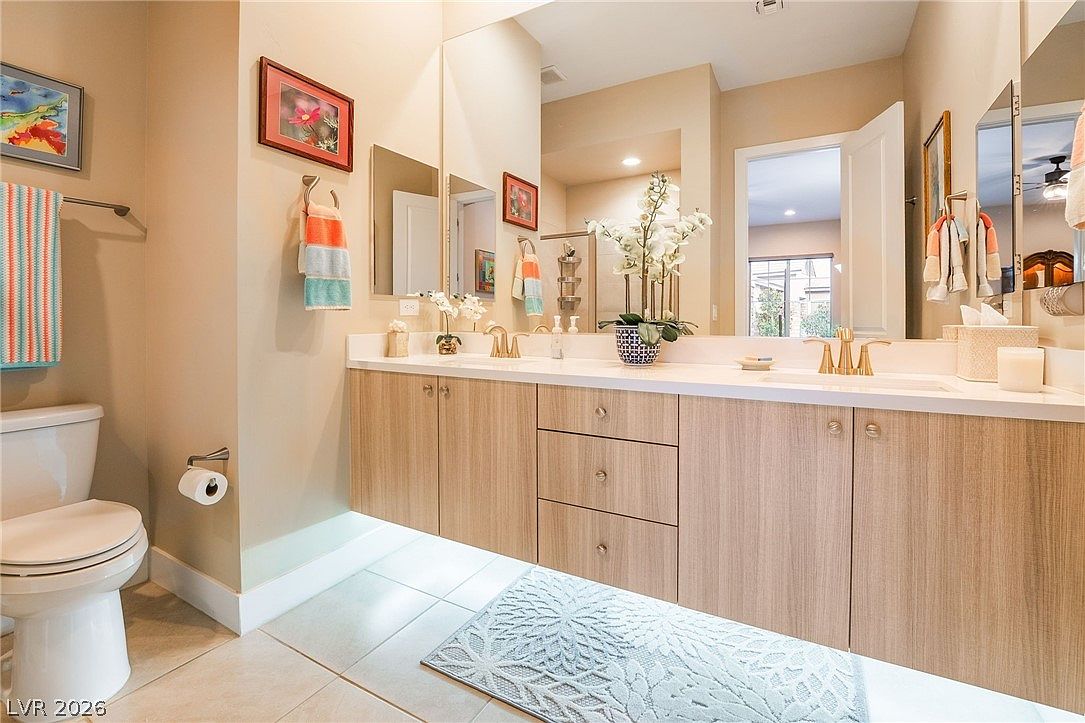 This bathroom features a contemporary floating vanity with light wood-tone cabinetry, brushed gold hardware, and a clean white countertop. The space is bright and airy, highlighted by a large wall-to-wall mirror, under-cabinet accent lighting, and decorative touches like a floral centerpiece and colorful towels. The overall impression is one of modern, functional style in a well-maintained guest or secondary bathroom.