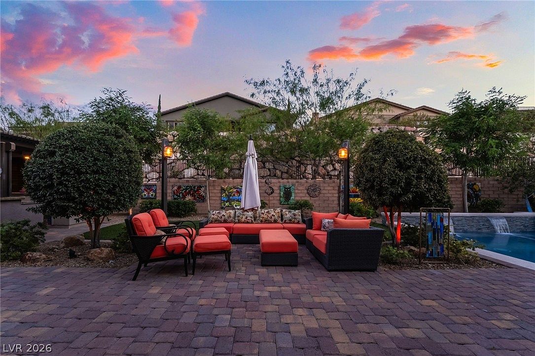 This inviting outdoor living space features a paved patio furnished with stylish, orange-cushioned wicker seating and an umbrella. The area is tastefully landscaped with shaped trees and wall-mounted art, positioned conveniently next to a tranquil swimming pool. The sunset lighting enhances the warm, relaxing atmosphere, making it a perfect spot for entertaining.
