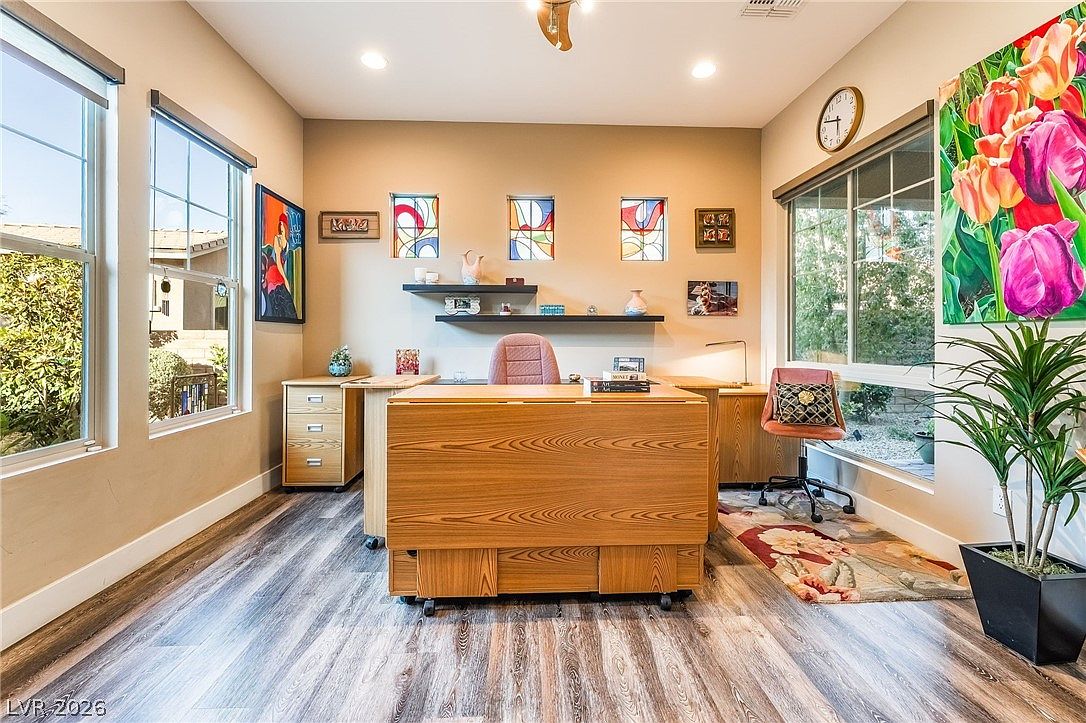 This bright and functional home office features light wood flooring and neutral walls, providing a clean canvas for productivity. The room includes a central desk setup, two floating shelves, and large windows that invite abundant natural light, while vibrant artwork and a potted plant add a touch of personality to the space.