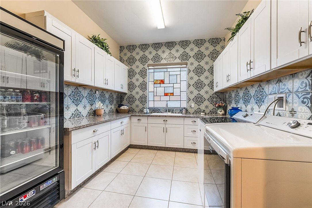 This functional and well-organized laundry room features white cabinetry, granite countertops, and a striking patterned tile backsplash that adds character to the space. It is equipped with a refrigerator and laundry appliances, creating a multi-purpose utility area. The room is brightened by an artistic stained-glass window above the sink, providing a unique architectural detail.