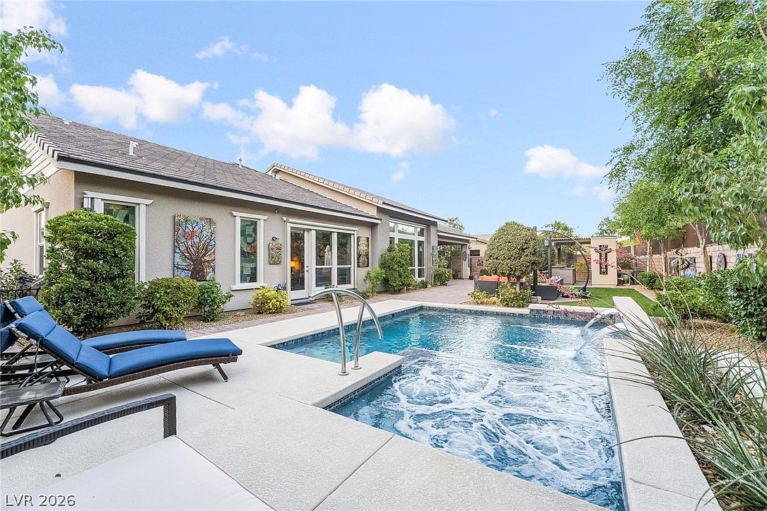 This outdoor space features a pristine swimming pool and integrated spa, perfect for relaxation and entertainment. The surrounding light-colored concrete deck, comfortable lounge chairs, and manicured landscaping create an inviting, modern resort-style atmosphere. The backyard seamlessly connects to the main residence through a sliding glass door, offering a harmonious blend of indoor-outdoor living.