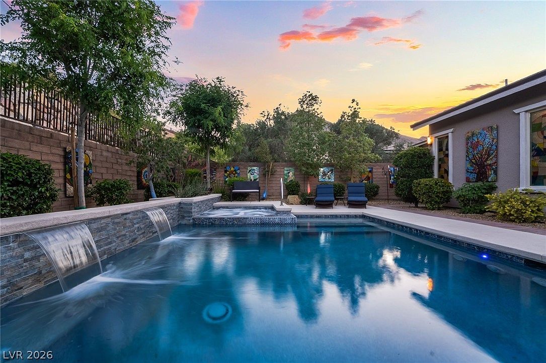 This stunning backyard features a pristine swimming pool with integrated water features and a raised spa, perfect for relaxation and entertaining. The surrounding area includes modern patio furniture, neatly landscaped greenery, and decorative artwork mounted on the perimeter wall. The scene is beautifully captured at sunset, highlighting a tranquil and inviting atmosphere.