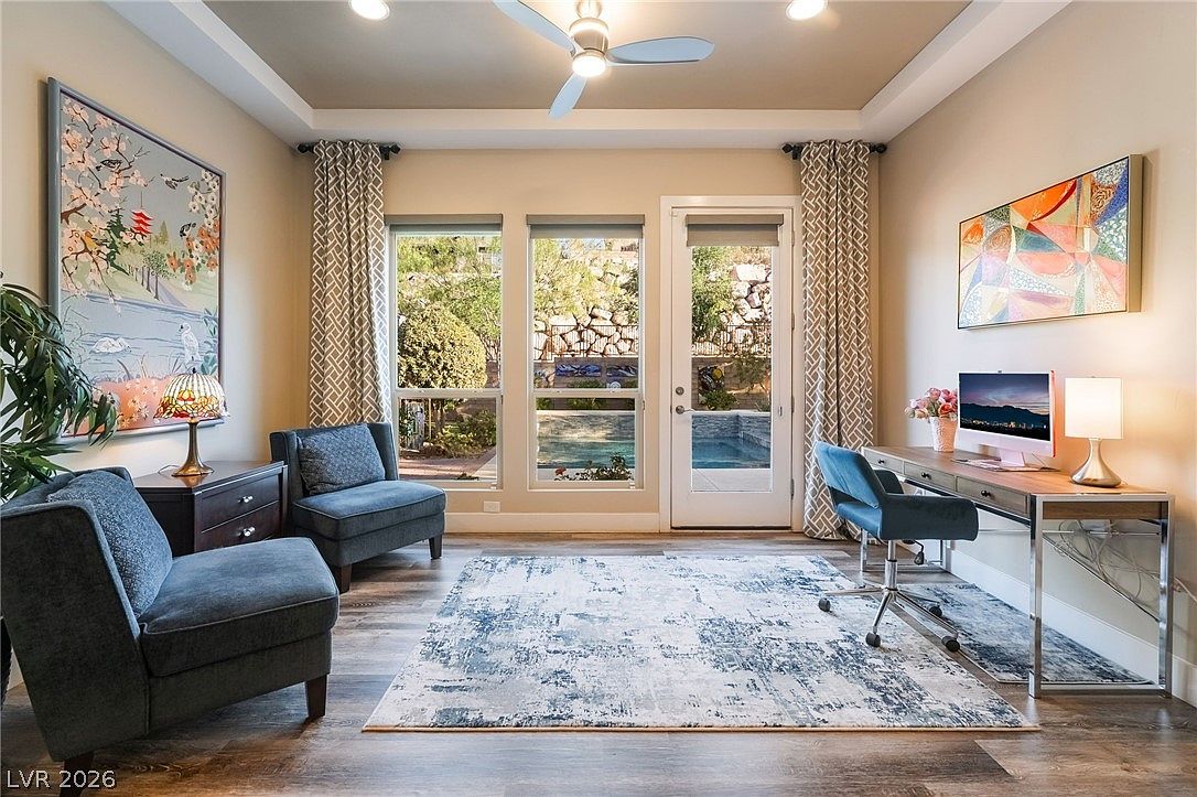 This bright and functional home office features wood-look flooring, a modern desk setup with an ergonomic chair, and comfortable seating for relaxation. Large windows and a glass-paned door provide natural light and a pleasant view of the outdoor pool area. The space is well-appointed with stylish wall art, patterned curtains, and a contemporary area rug, creating an inviting and professional atmosphere.