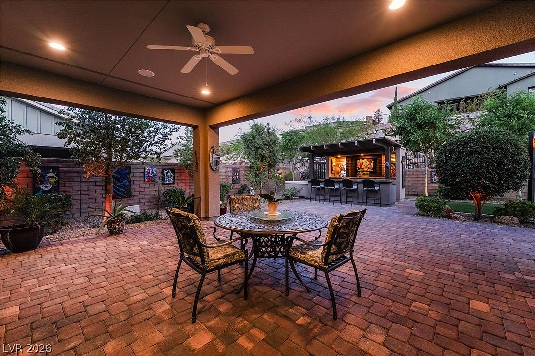 This inviting covered patio features a stamped brick paver floor and a comfortable outdoor dining set, perfect for alfresco meals. The space includes a ceiling fan for climate control and overlooks a professionally landscaped backyard with a built-in outdoor bar area, creating an ideal setting for entertaining. The warm lighting and cohesive design elements offer a relaxed, upscale atmosphere.