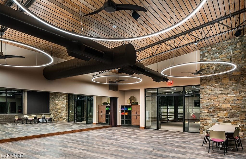 This spacious community activity room features a modern, industrial-inspired aesthetic with high wooden ceilings, circular pendant lighting, and exposed ductwork. The room includes a wall of mirrors, built-in shelving for storage, and a designated dining area, making it ideal for group classes or social events. Large glass doors and stone accent walls add a sophisticated, welcoming touch to this functional shared space.