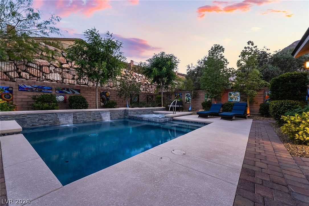 This stunning backyard features a pristine, rectangular swimming pool with integrated water features and a raised spa. The patio area is elegantly paved with light-toned concrete and stone accents, complemented by two lounge chairs and decorative wall art. Surrounded by mature trees and a scenic rock wall, the space offers a tranquil and high-end atmosphere perfect for outdoor relaxation.