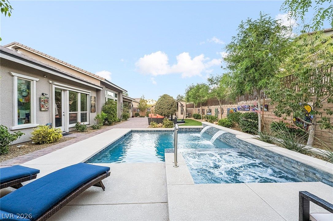 This outdoor space features a modern, rectangular swimming pool equipped with integrated water features and a raised spa area with jets. The surrounding patio is constructed from light-toned concrete, complemented by well-maintained landscaping, trees, and decorative artwork on the wall. This area serves as a luxurious, private backyard retreat ideal for relaxation and entertaining.