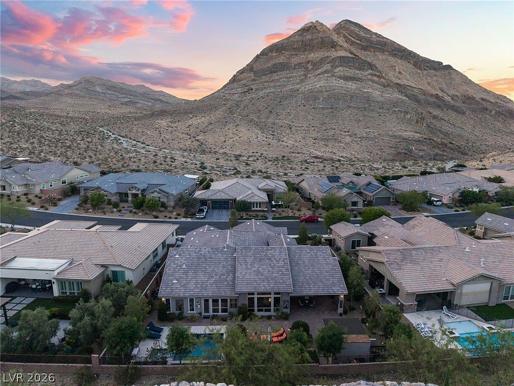 This stunning aerial view highlights a residential neighborhood nestled at the base of a majestic, craggy mountain range. The focal property features a backyard with a private swimming pool, tiled roof, and a seamless transition to the outdoor living area. The desert-inspired landscaping and sunset lighting create an inviting and tranquil atmosphere.