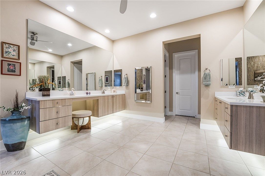 This spacious primary bathroom features a double-vanity layout with modern wood-grain cabinetry and crisp white countertops. The room is accented by large, polished mirrors, neutral tile flooring, and sophisticated recessed lighting, creating a bright and contemporary atmosphere. A dedicated vanity seating area and a clean, minimalist design define this functional and elegant space.