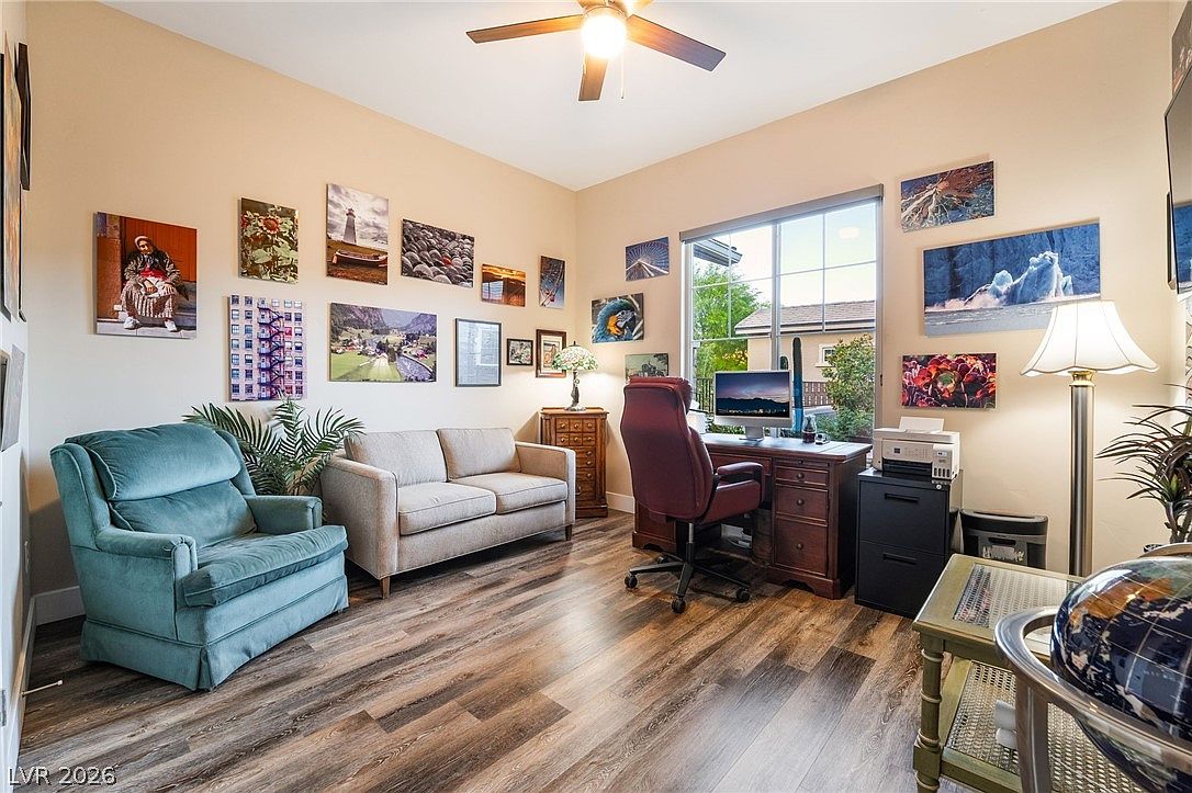 This home office features wood-look plank flooring, a dark wooden desk with a matching leather executive chair, and a cozy seating area with a light-toned sofa and a teal armchair. The room is decorated with a gallery wall of landscape photography, and a large window provides natural light, creating a professional yet comfortable workspace.