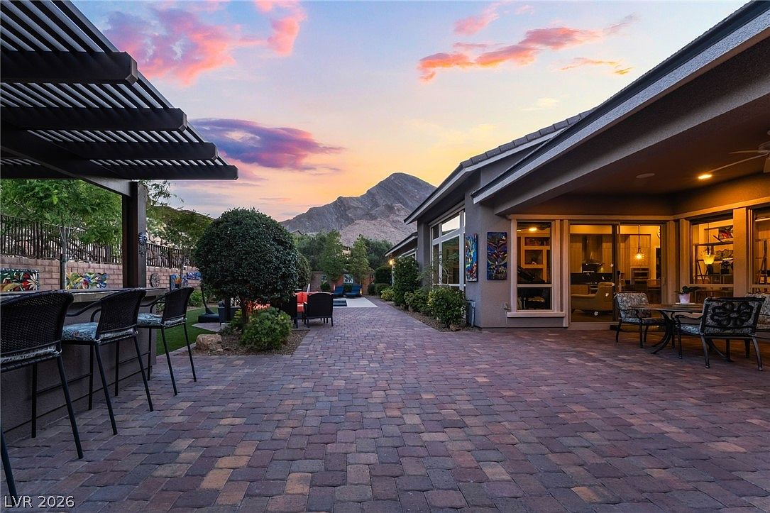 This spacious outdoor patio features beautiful paver stones and an integrated bar area with bar stool seating, perfect for entertaining. The covered living area looks out over a well-manicured yard and stunning mountain views, all set against a vibrant sunset sky. This space offers a seamless transition between indoor and outdoor living, highlighting a tranquil and luxurious desert lifestyle.