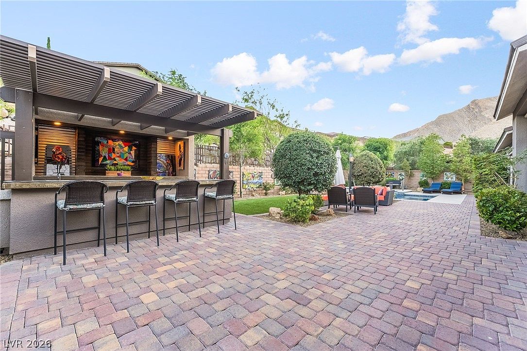 This spacious backyard features a covered outdoor kitchen and bar area equipped with high-top seating, perfect for entertaining. The patio is laid with durable, multi-toned pavers that lead toward a seating area and an inviting swimming pool. The desert landscape, complete with groomed shrubs and a scenic mountain backdrop, creates a beautiful and relaxing outdoor retreat.