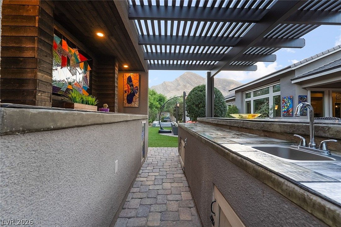 This outdoor patio space features a fully equipped kitchenette complete with a sink and counter space, sheltered by a modern pergola. The area is tastefully decorated with colorful wall art and overlooks a landscaped backyard with a mountain view in the distance. The combination of brick-style pavers and built-in amenities makes this an ideal space for outdoor entertaining.