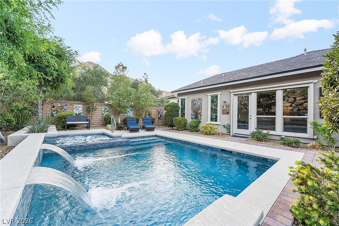 This appealing backyard features a modern swimming pool with a raised spillover spa and elegant water features. The area is complemented by stone decking, lounge chairs, and artfully decorated walls, creating a serene oasis with desert landscaping and mountain views in the background. It is a perfect space for outdoor relaxation and entertaining.
