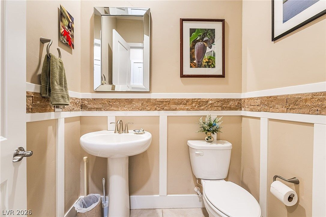 This compact powder room features a pedestal sink and a white toilet, complemented by a stone tile backsplash that adds texture to the space. The room is decorated with framed wall art and a simple mirror, creating a neat and inviting environment. Neutral wall tones and crisp white trim give the room a polished, classic aesthetic.