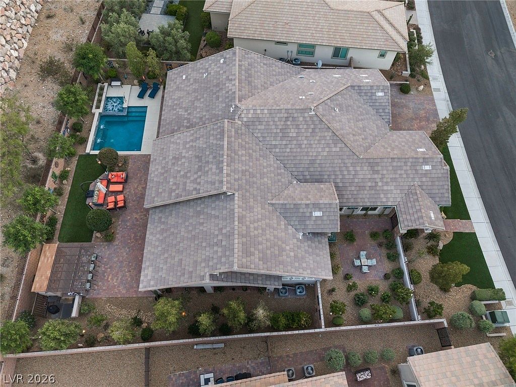 This aerial view showcases a well-maintained property featuring a large tiled roof, a private swimming pool with an integrated spa, and a spacious paver patio area perfect for outdoor entertaining. The landscaping includes manicured garden beds and a covered patio structure, all set within a private fenced yard adjacent to a suburban street. The overall impression is one of a comfortable, upscale residential property designed for both leisure and curb appeal.