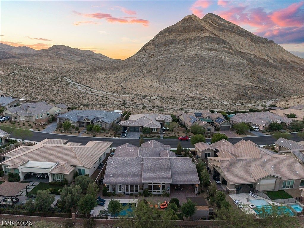 This aerial view showcases a desert residential neighborhood nestled against the base of a prominent mountain range during a scenic sunset. The properties feature modern suburban architecture with neutral tile roofing, landscaped backyards containing private swimming pools, and spacious layouts. The high-angle perspective highlights the proximity of the homes to expansive natural vistas and the surrounding arid landscape.