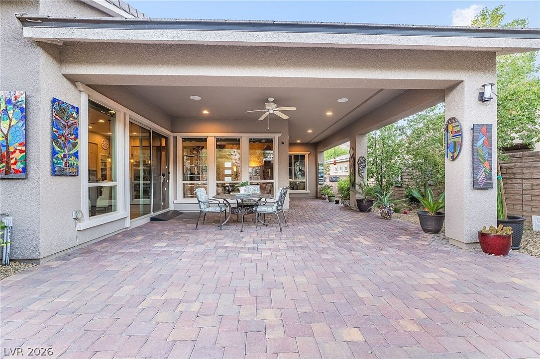This covered patio features classic brick-style pavers and provides a spacious outdoor living area with a built-in ceiling fan and recessed lighting. The space is adorned with colorful wall art and potted plants, creating a welcoming and comfortable setting for relaxation or entertaining. Large sliding glass doors offer a seamless transition between the indoor living area and the well-maintained backyard.