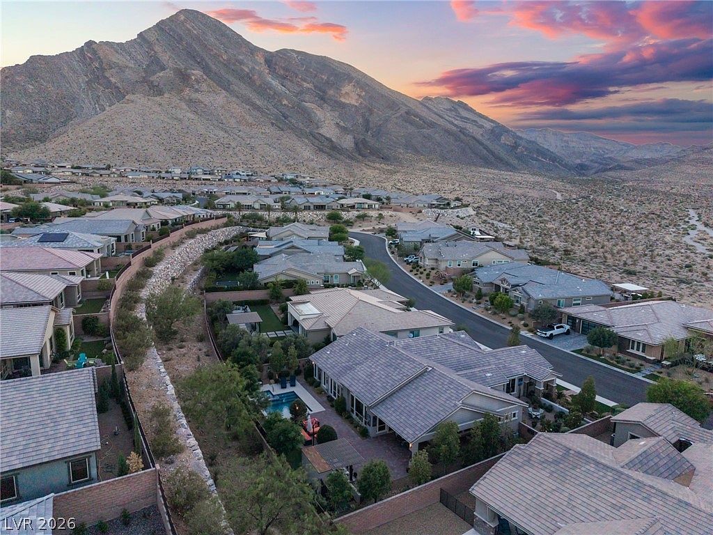 This high-angle aerial view showcases a modern suburban neighborhood nestled against a dramatic mountain range at sunset. The featured home includes a well-maintained backyard with a private swimming pool, patio area, and desert landscaping. The overall setting offers a picturesque blend of luxury residential living and stunning natural scenery.