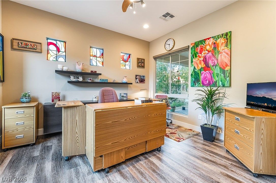 This home office features a wood-grain desk as its centerpiece, complemented by matching storage cabinets and modern wood-look laminate flooring. The space is brightened by unique stained-glass accent windows and a large piece of floral wall art, creating a cozy and functional work environment. A large window provides natural light and a pleasant view, making it an inviting space for productivity.