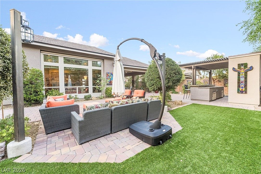 This spacious backyard patio features a cozy conversational seating area with grey wicker furniture and orange cushions, anchored by a large, adjustable cantilever umbrella. Adjacent to the seating area is an outdoor kitchen setup with a bar area, all set against a well-maintained lawn and paved brick flooring. The space provides a serene and inviting environment for outdoor dining and relaxation.