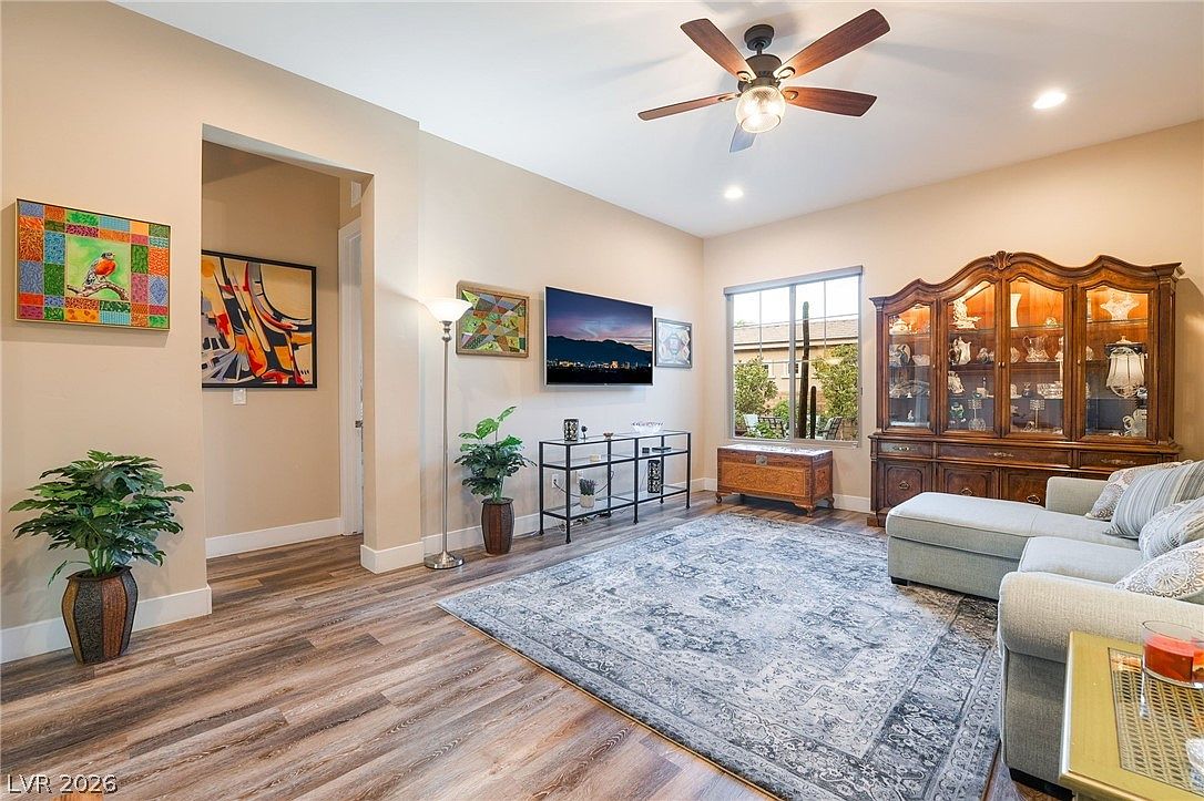 This inviting living room features a spacious, open-concept layout with wood-style flooring and neutral wall tones. Key highlights include a mounted television above a minimalist metal display table, a traditional wooden china cabinet, and a comfortable sectional sofa accented by an ornate, large-scale area rug. Large windows provide natural light, while vibrant abstract artwork adds a personalized, colorful touch to the space.