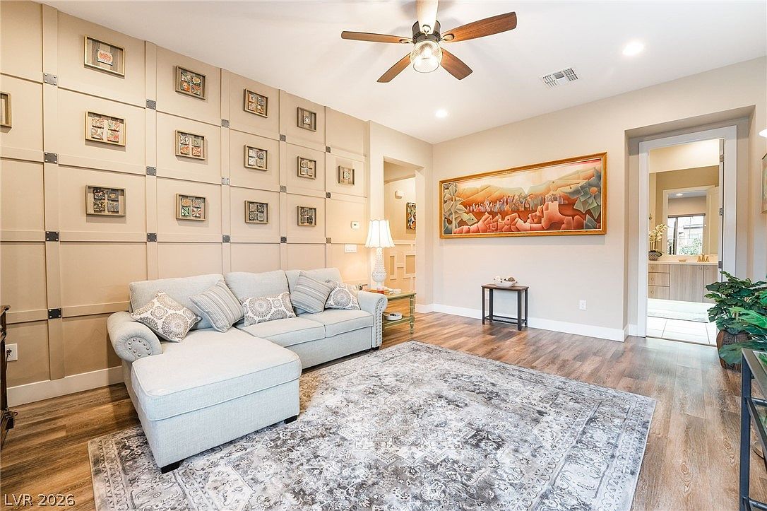 This living room features a contemporary design highlighted by an expansive, grid-paneled feature wall adorned with multiple small framed artworks. The space includes a comfortable light-colored sectional sofa, a textured patterned rug, and a large, vibrant landscape painting that serves as a focal point. Warm wood-look flooring and a ceiling fan complete this inviting and well-lit residential living area.