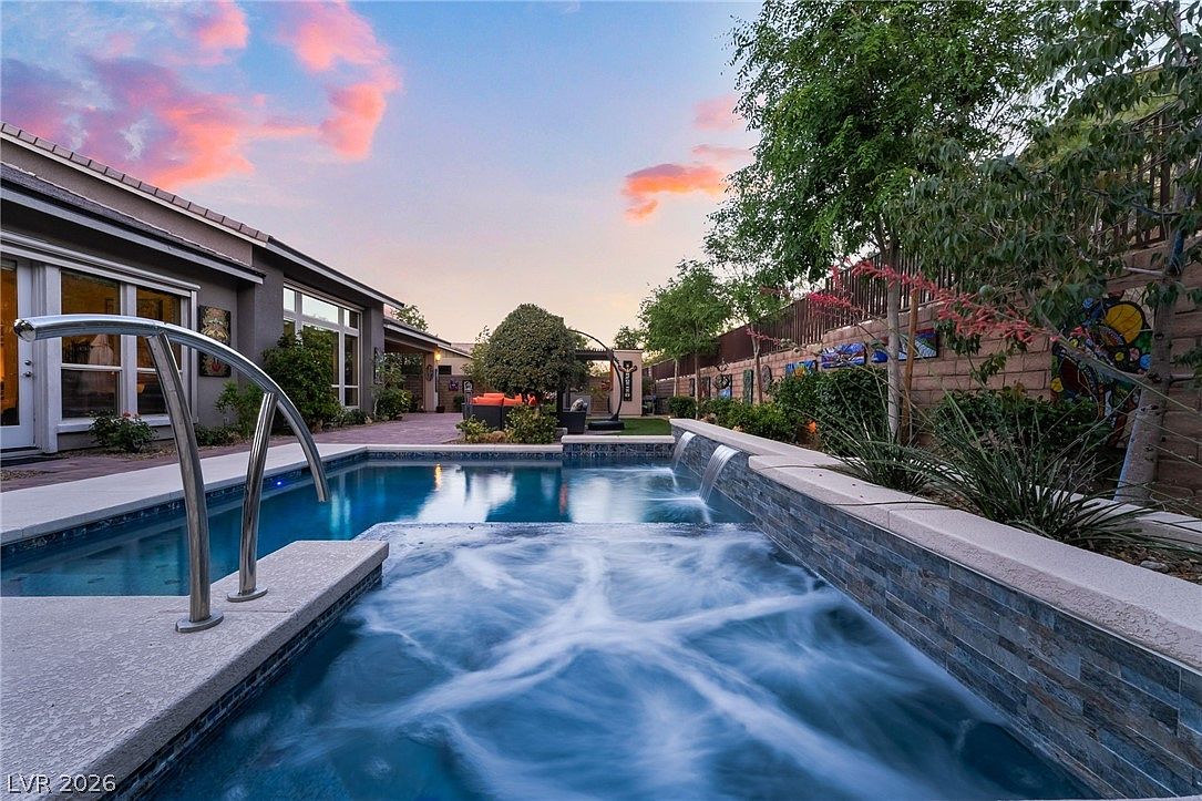 This stunning backyard oasis features a beautiful pool and integrated spa, highlighted by a contemporary stone water feature that cascades into the water. The serene setting includes a patio area with comfortable seating and thoughtful landscaping, perfect for relaxation and outdoor entertaining. The twilight sky creates a warm, inviting atmosphere for this high-end residential outdoor space.