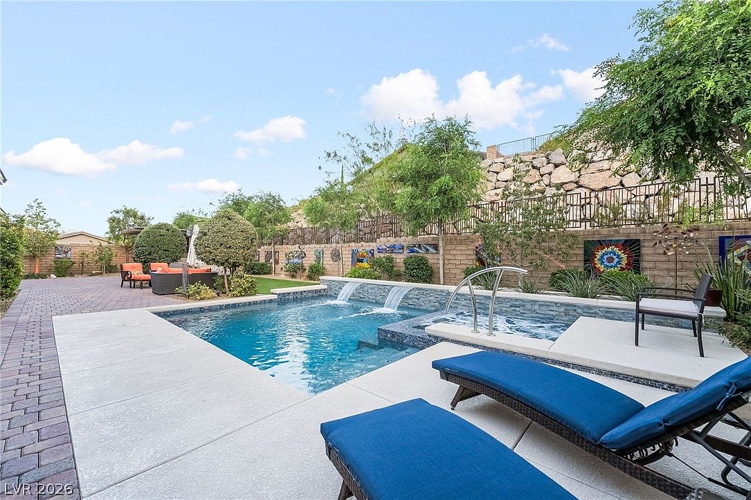 This backyard features a modern swimming pool with an integrated spa, complete with two waterfall features and tile accents. The surrounding area includes light-colored concrete decking, two lounge chairs, and a paved patio area with outdoor seating. The space is landscaped with various shrubs and trees, framed by a decorative stone retaining wall.
