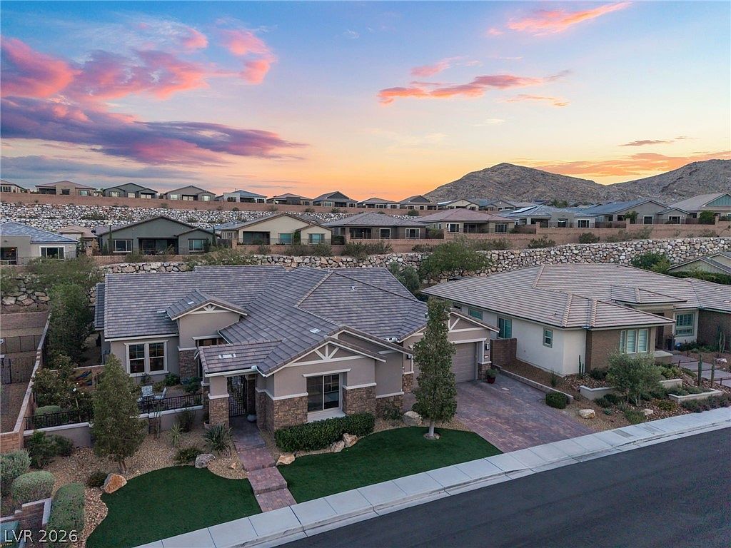 This elevated view captures a well-maintained single-story suburban residence featuring a neutral color palette with stone veneer accents and a tiled roof. The property boasts a manicured lawn, a paved driveway, and an inviting entry walkway, situated within a modern residential development with a scenic desert mountain backdrop. The overall aesthetic is clean and contemporary, typical of planned communities in arid regions.