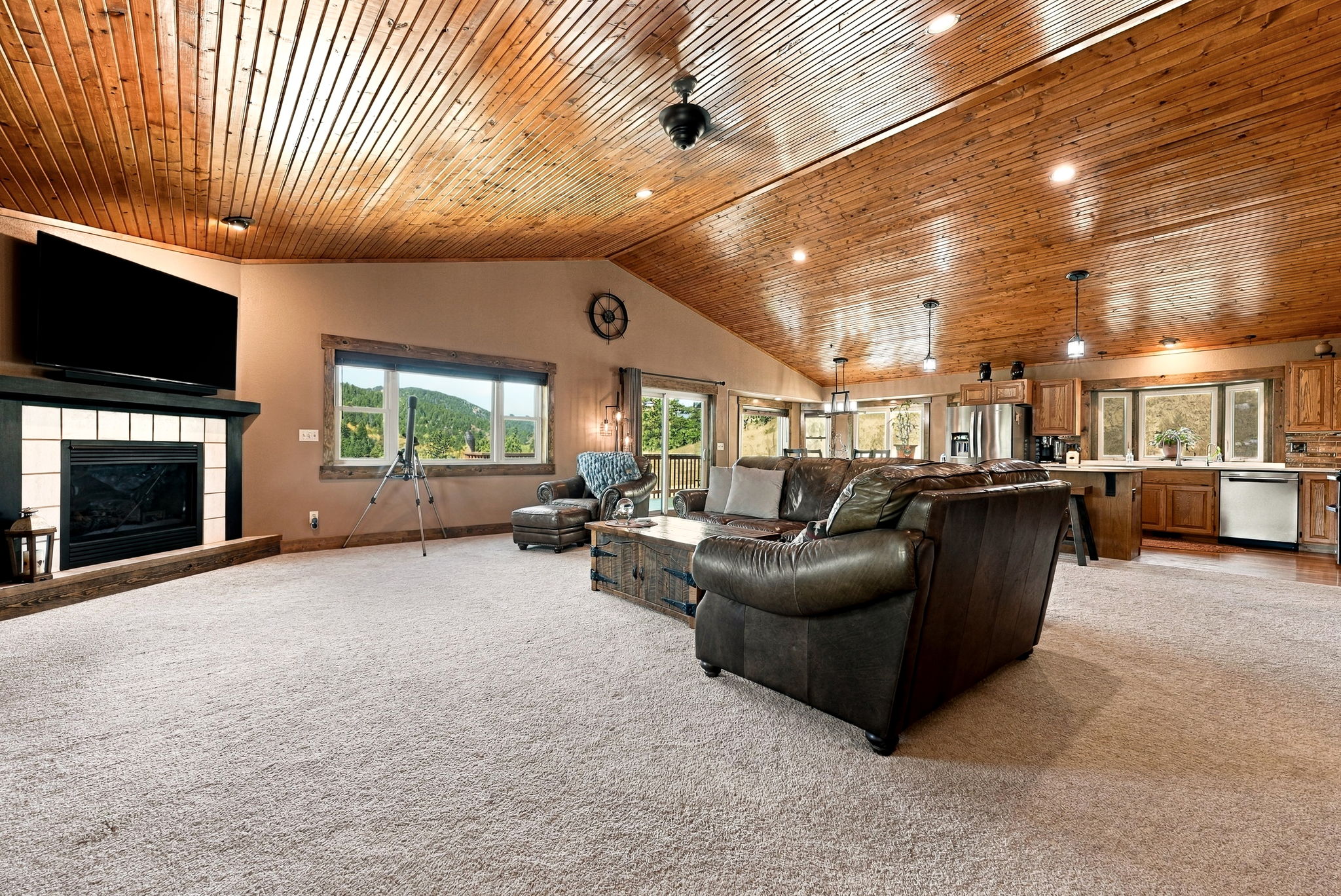 This spacious open-concept living area features a vaulted wood-paneled ceiling and neutral carpeting throughout. A large leather sectional sofa faces a fireplace with a light-colored tile surround, while a nearby window offers a scenic view of the surrounding hills. The space flows seamlessly into a kitchen area in the background, creating an inviting and airy atmosphere.