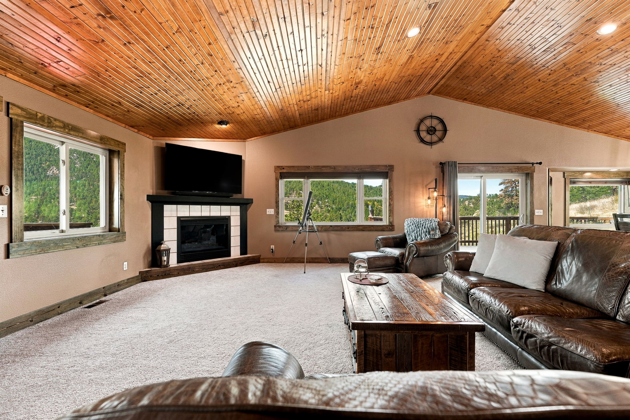 This spacious living room features a vaulted wood-paneled ceiling and a cozy fireplace with a dark mantel and light-colored tile surround. The room is furnished with a large leather sofa and a matching armchair, centered around a rustic wooden coffee table. Large windows provide ample natural light and scenic views of the surrounding forest, creating a warm and inviting atmosphere.