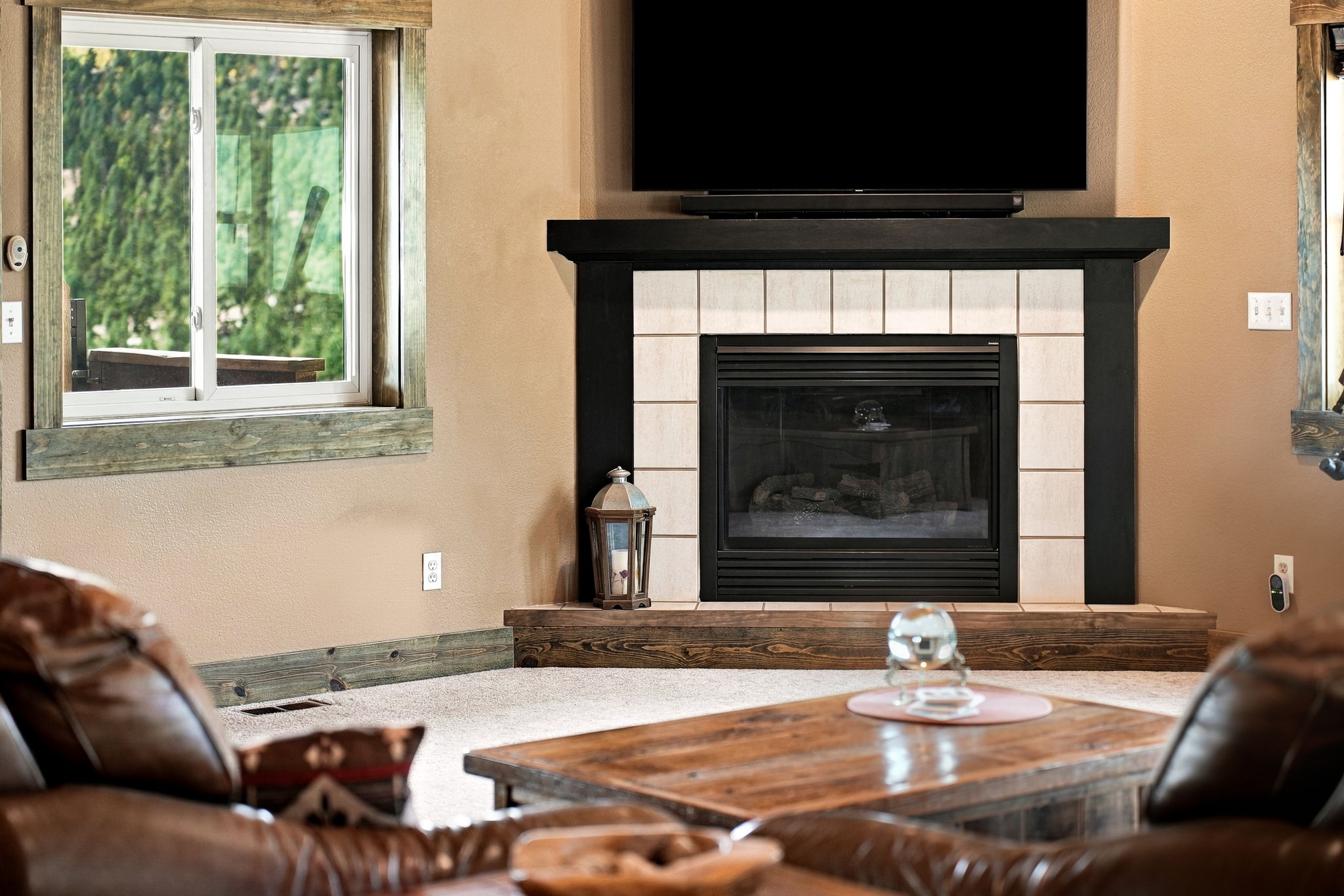 This cozy living room features a corner fireplace with a black frame and a light-colored tile surround, topped by a wall-mounted television. A rustic wooden mantel and hearth anchor the fireplace, while a large window to the left provides natural light and a view of the outdoors. The foreground includes a wooden coffee table and leather seating, creating a warm and inviting atmosphere.